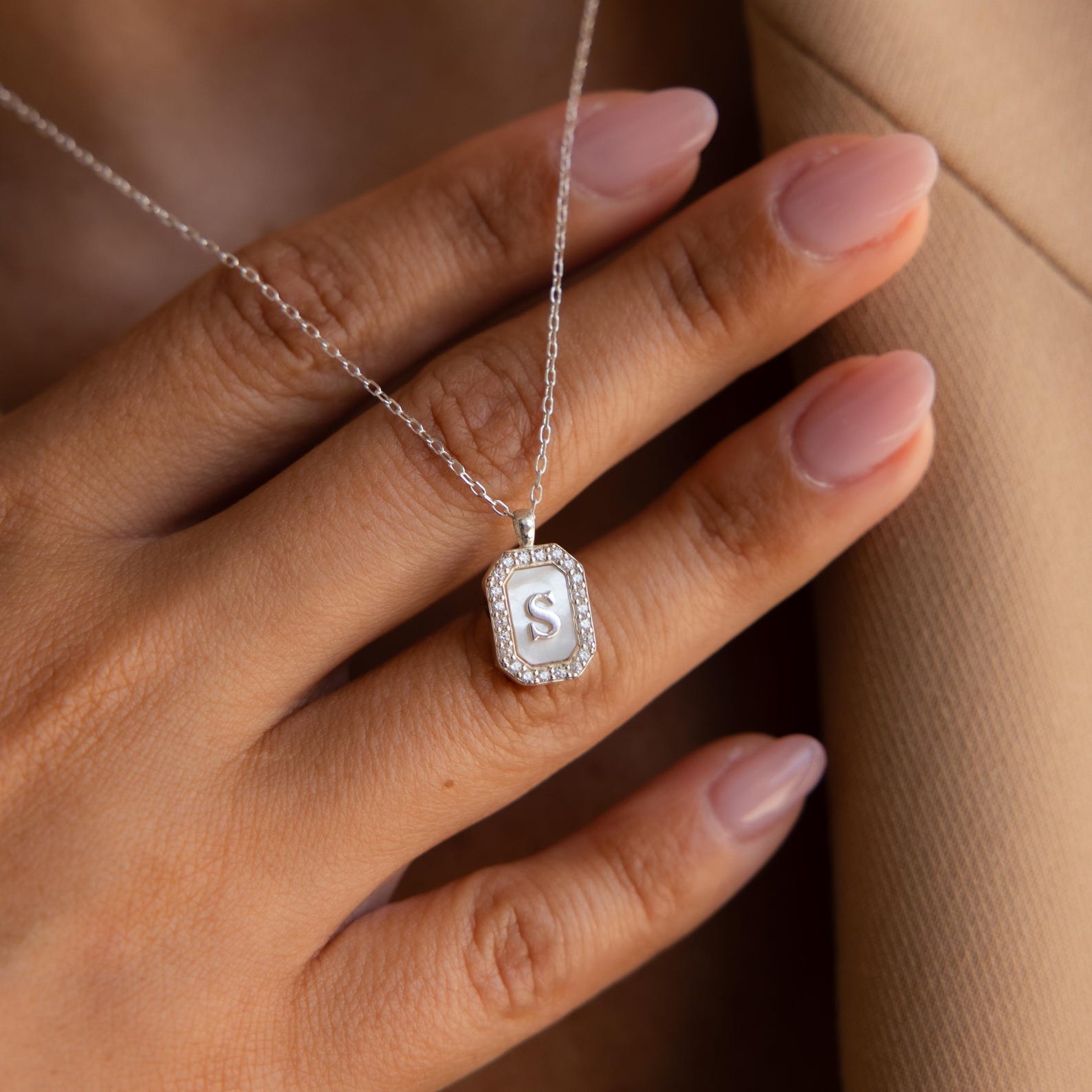 A hand with pale pink nails holds the Pave Gemstone Initial Necklace, which features an S-shaped pendant accented by sparkling gemstones.