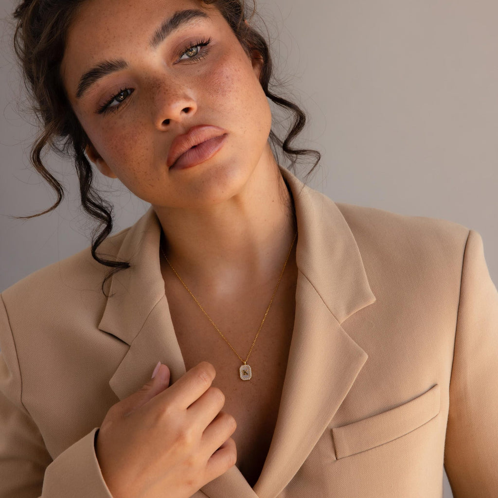 A woman in a beige blazer wears the Pave Gemstone Initial Necklace, looking slightly away with a neutral expression.