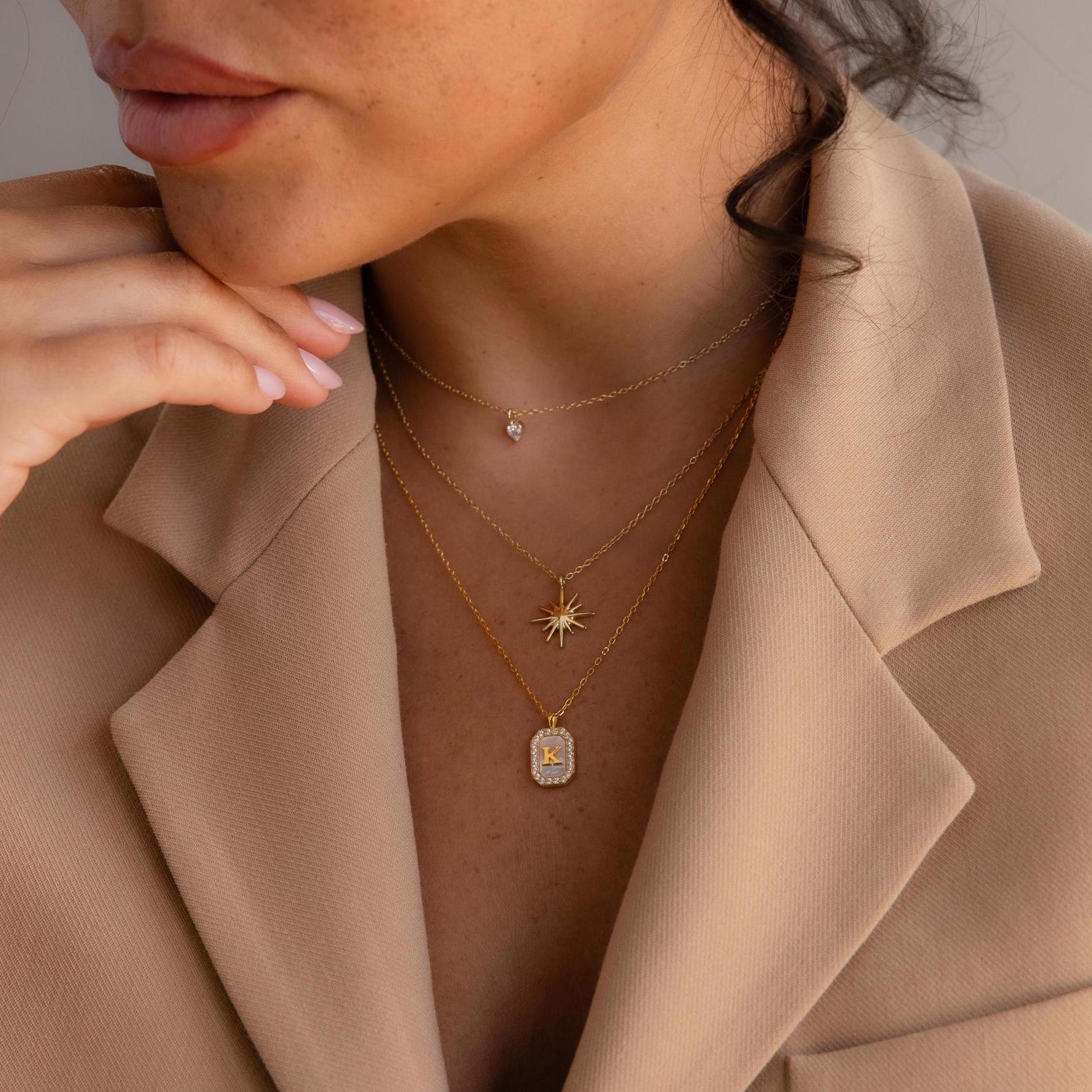 Woman wearing a beige blazer and layered gold necklaces, including a Pavé Gemstone Initial Necklace featuring the letter K.