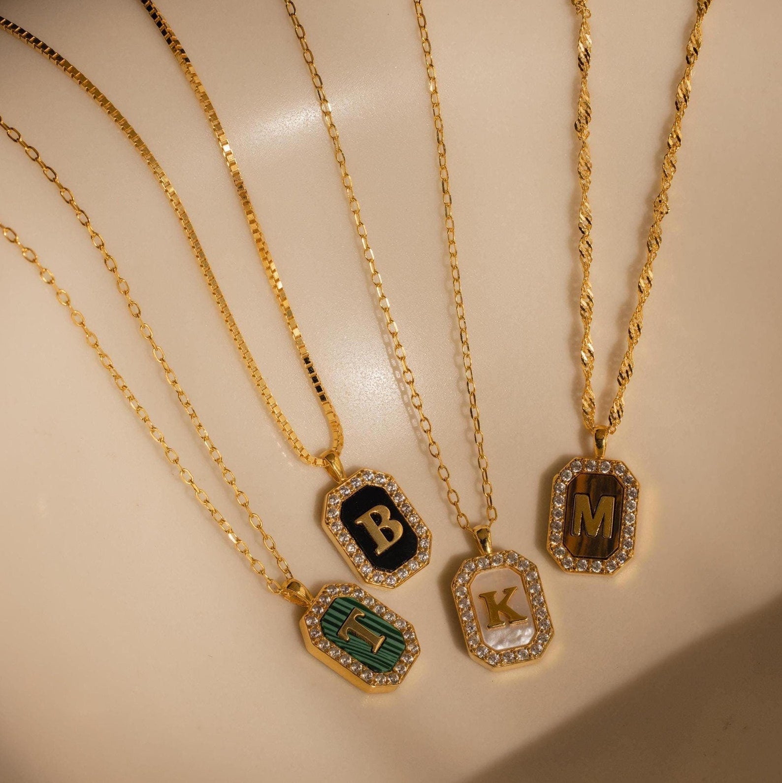 Five Pave Gemstone Initial Necklaces with gold letter pendants B, M, L, K, and E feature delicate gemstone inlays and are displayed on a light beige surface.