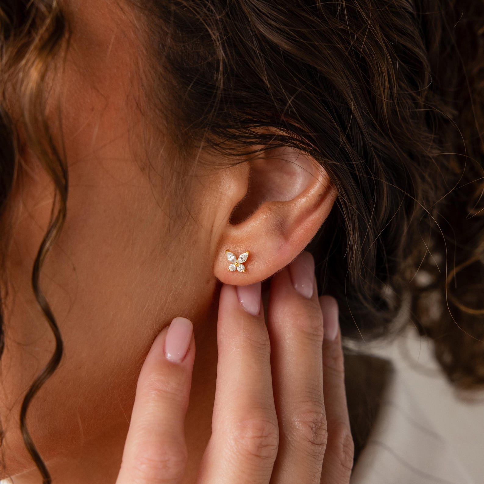 A woman touches her ear, wearing Butterfly Birthstone Studs—elegant gold butterfly earrings that add a hint of sparkle and charm.