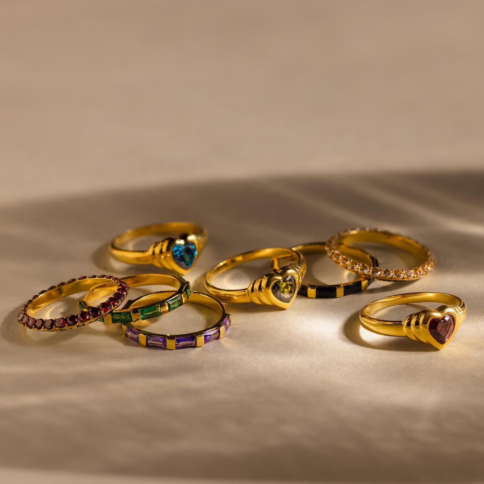 Eight Half Eternity Birthstone Rings are arranged on a beige surface with soft shadows—an elegant choice for an anniversary keepsake.