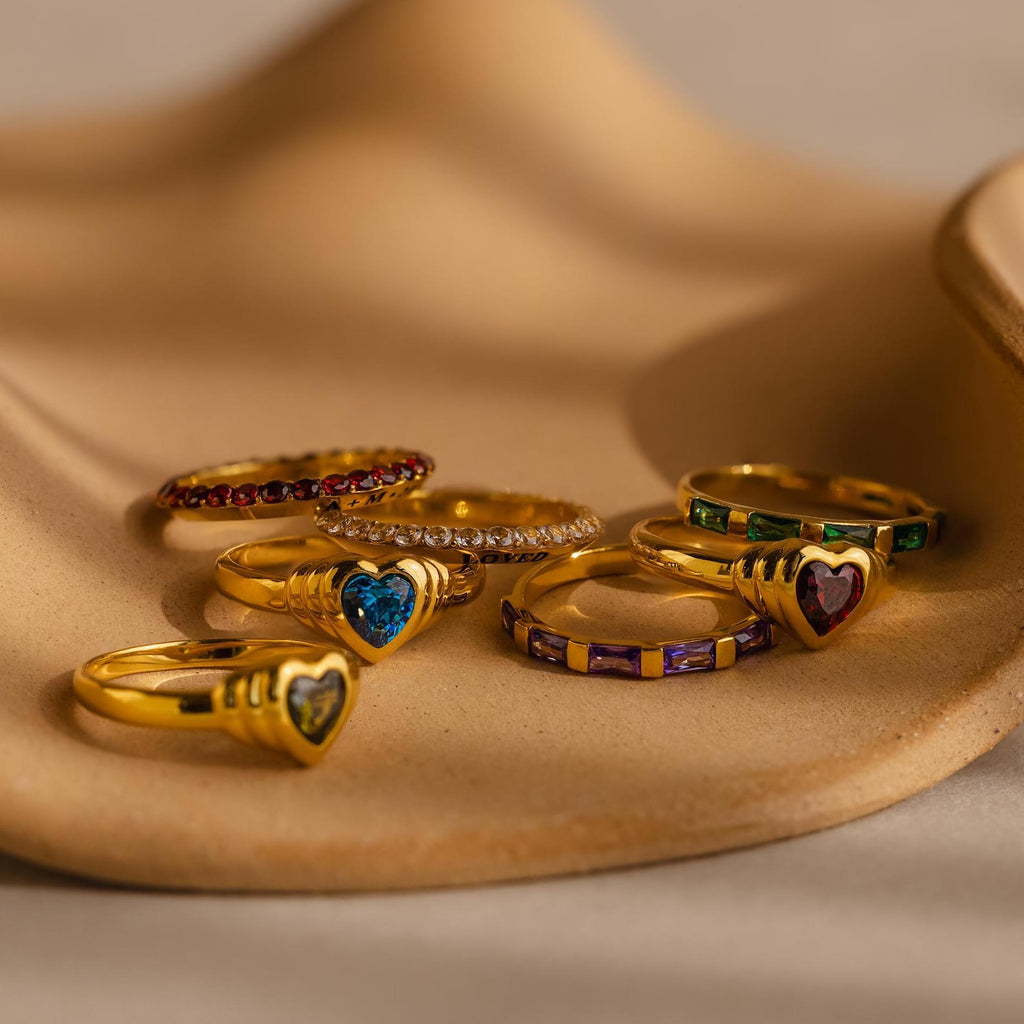 Six gold rings with vibrant gemstones and heart designs—featuring a personalized engraved ring and the Engraved Birthstone Eternity Ring—are displayed on a beige, wavy surface.