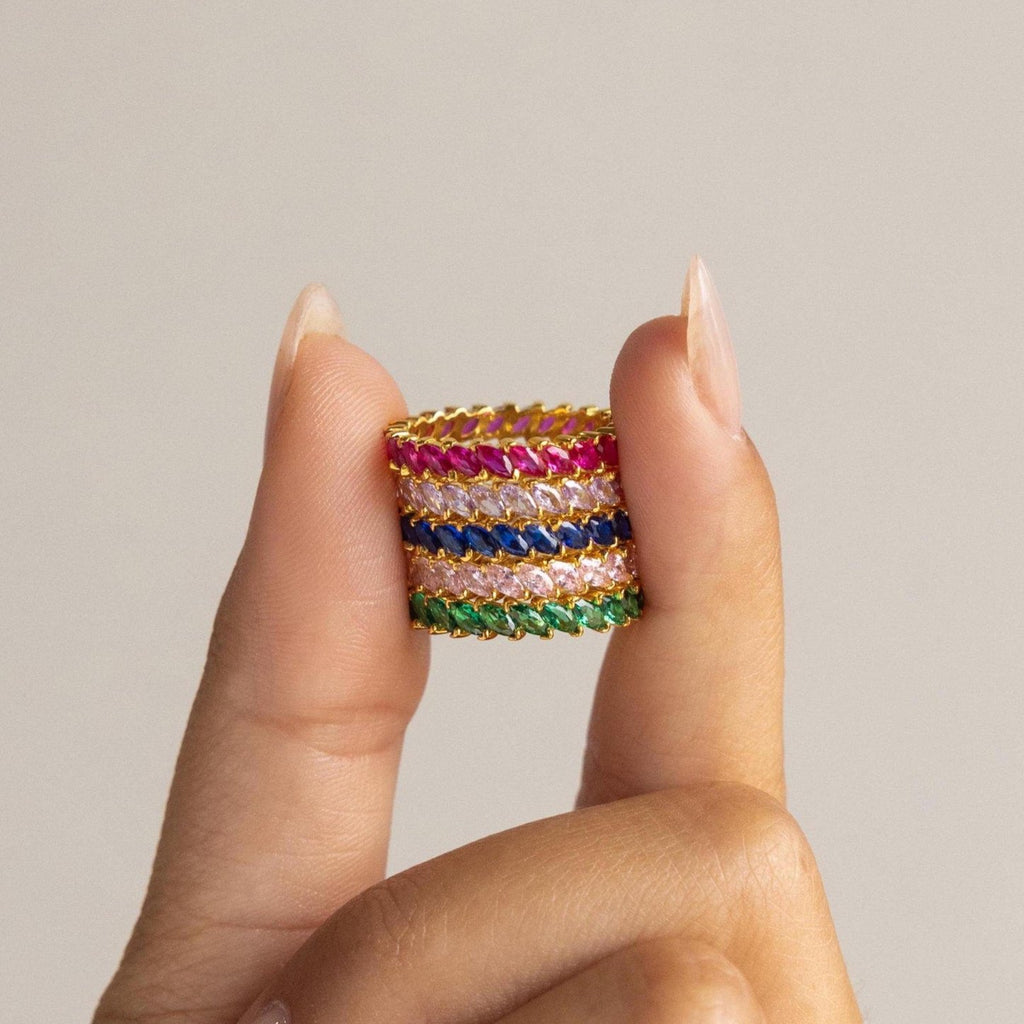 A hand displays the Eternity Marquise Birthstone Ring—four gold bands with pink, red, blue, and green marquise stones stacked between two fingers—an elegant customizable piece, ideal as a unique Christmas gift.