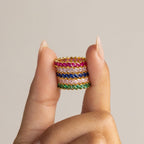 A hand displays the Eternity Marquise Birthstone Ring—four gold bands with pink, red, blue, and green marquise stones stacked between two fingers—an elegant customizable piece, ideal as a unique Christmas gift.