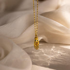 The Evil Eye Birthstone Hand Necklace features a hand-shaped gold pendant on a chain, shown against light textured fabric with gentle lighting.