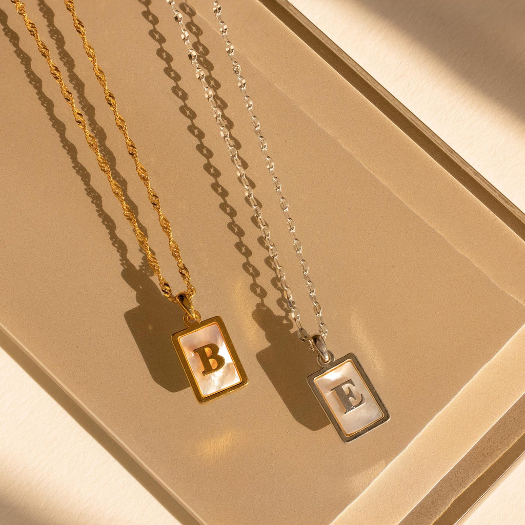 Two Pearl Initial Tag Necklaces with rectangular pendants—one gold with a B, one silver with an E—rest on a beige surface. Personalize yours with your chosen letter and chain, making it a perfect Christmas gift.