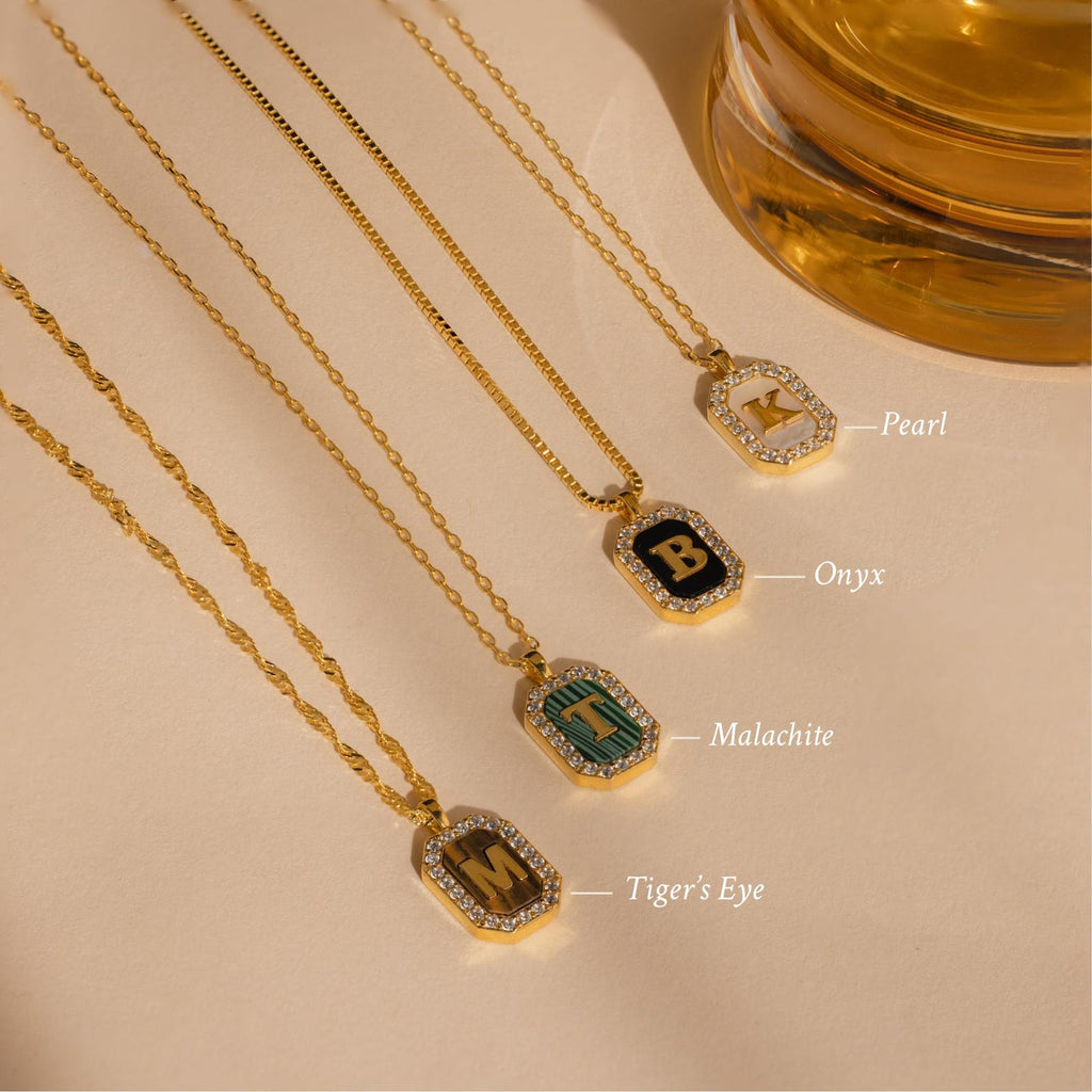 Four Pave Gemstone Initial Necklaces feature gold letter pendants inlaid with pearl, onyx, malachite, and tiger’s eye gemstones, displayed near a glass.