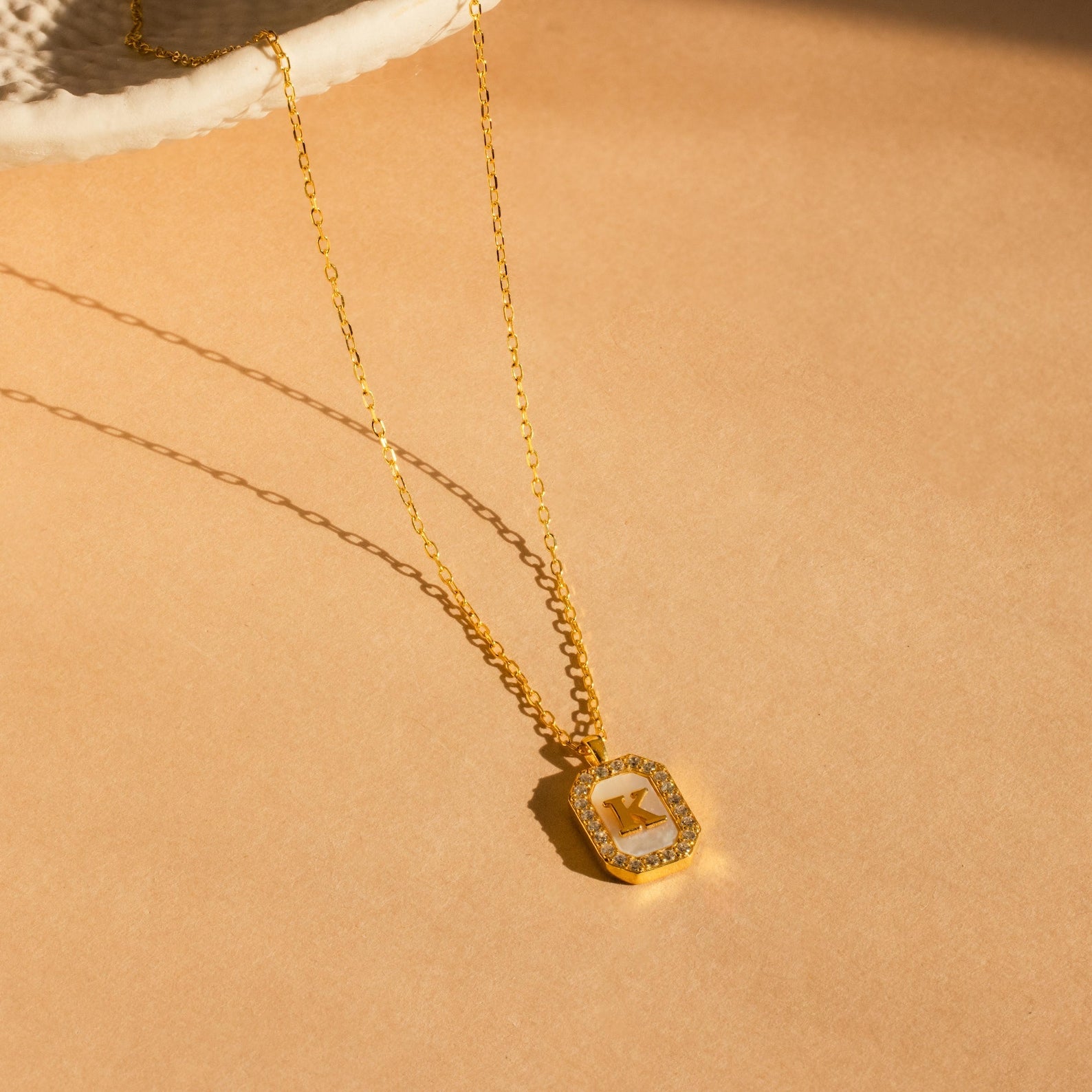 The Pave Gemstone Initial Necklace, featuring a rectangular K pendant with sparkling stones, is displayed on a beige surface in sunlight.