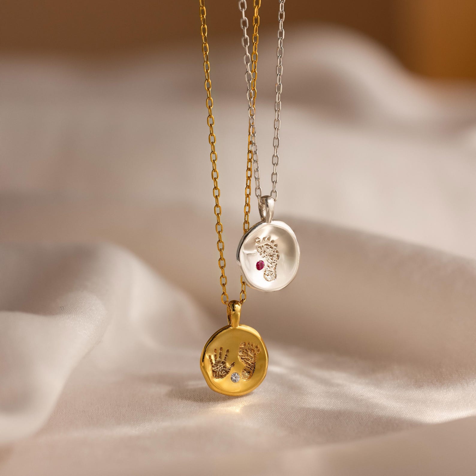 The Baby Handprint Coin Necklace, a gold or silver pendant engraved with a precious print, rests on soft cream fabric—an ideal personalized keepsake and thoughtful new mom gift.