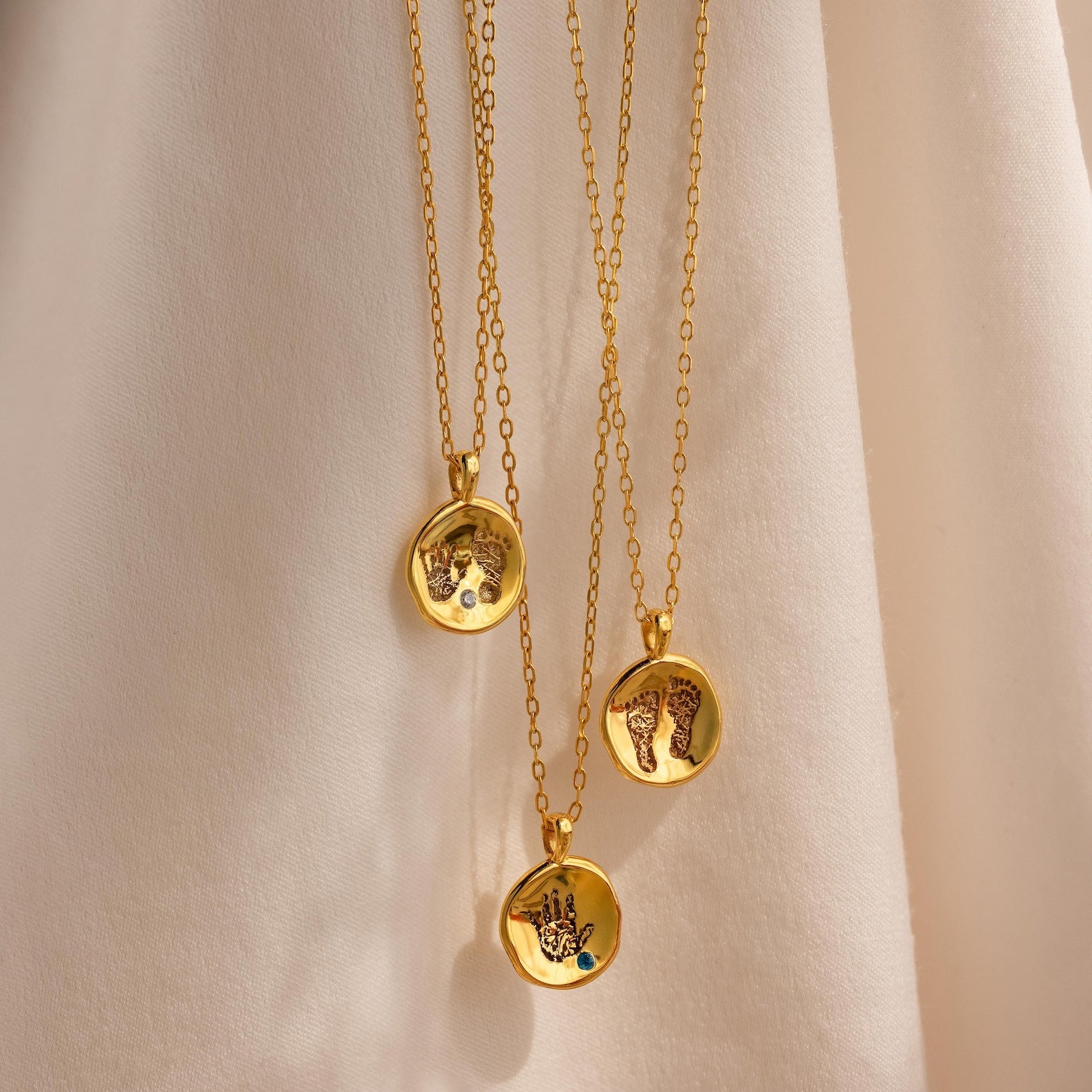The Baby Handprint Coin Necklace features three gold pendants engraved with hand and foot designs, displayed on soft beige fabric—a personalized keepsake and thoughtful gift for new moms.