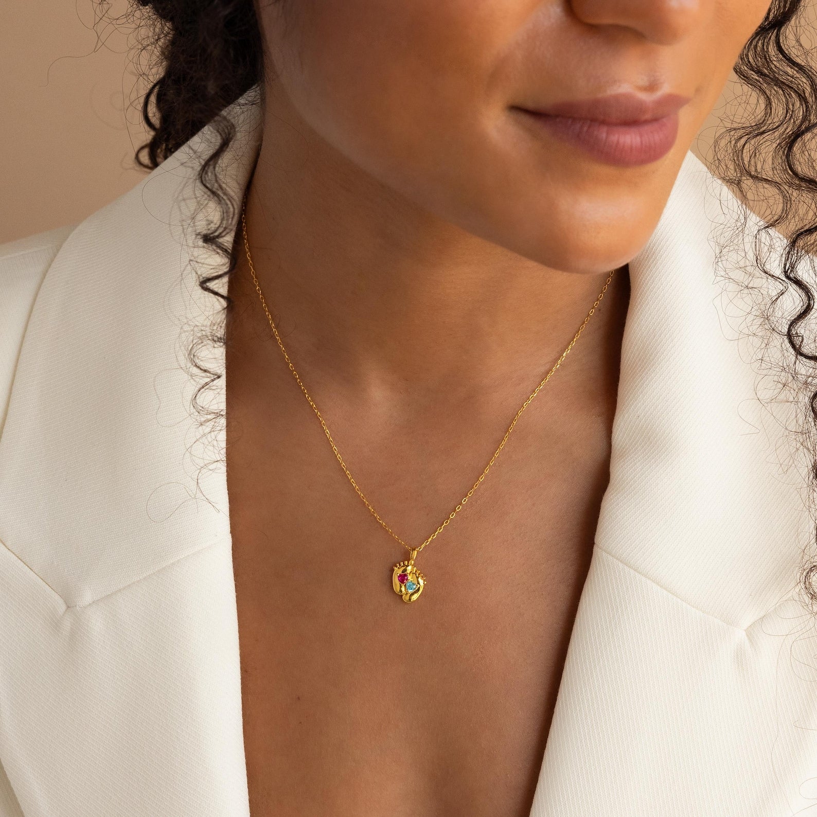 Woman in a white blazer wearing the Footprint Birthstone Necklace—a personalized charm necklace with a colorful birthstone pendant, shown from the shoulders up.