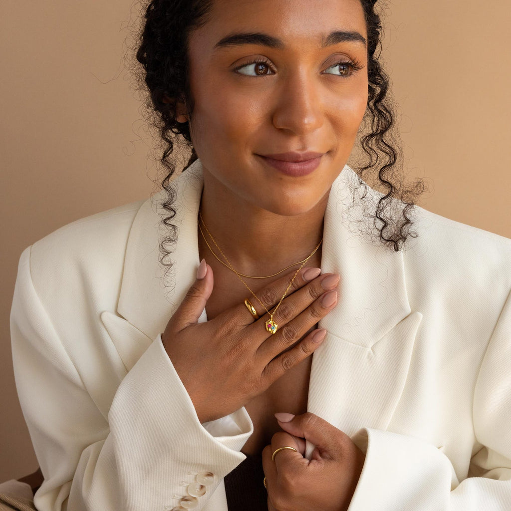 Woman in a white blazer smiles softly, touching her gold rings and Footprint Birthstone Necklace, with a beige background.