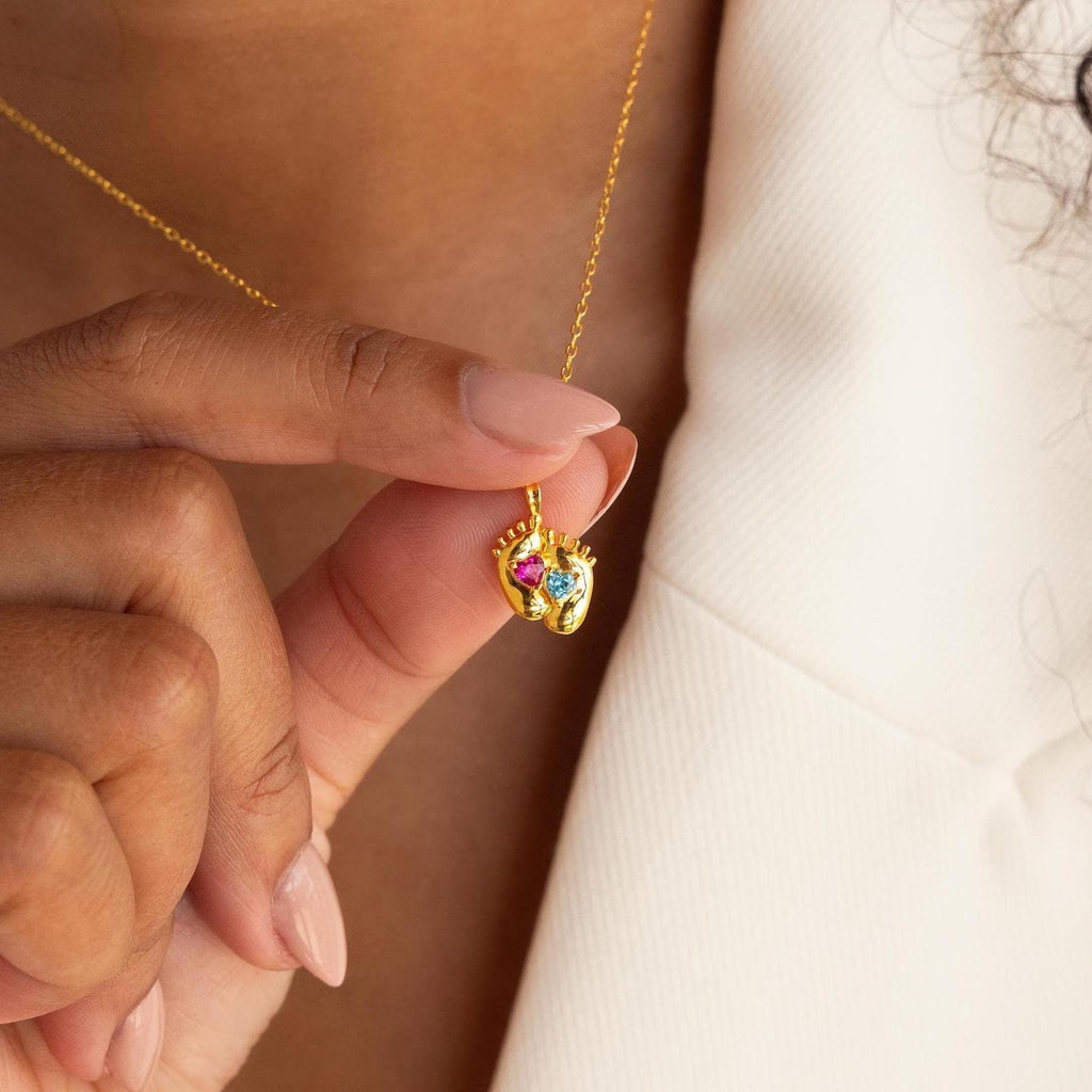 A person holds the Footprint Birthstone Necklace, a gold personalized charm necklace featuring colorful birthstone accents and small sculpted hands.