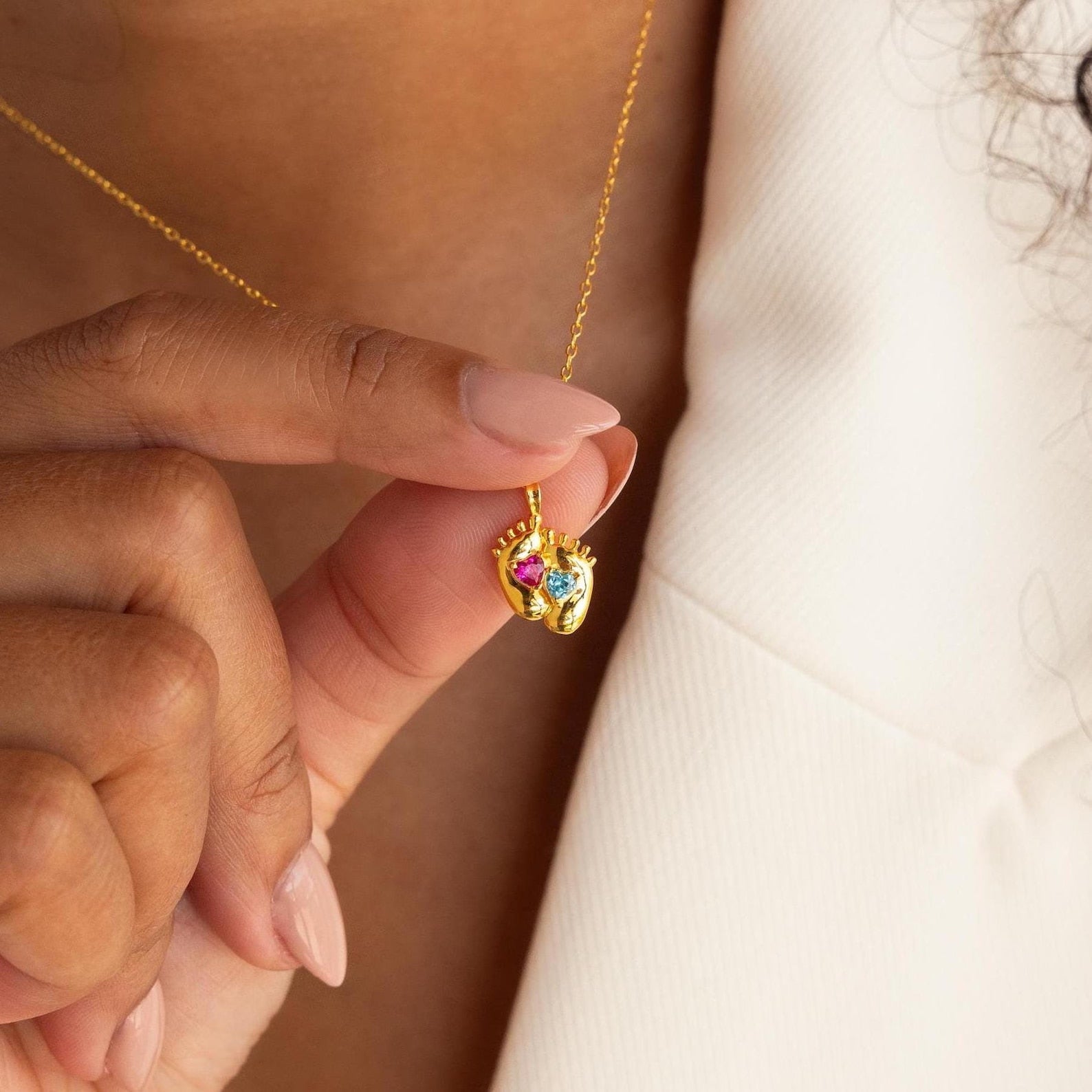 A person holds the Footprint Birthstone Necklace, a gold personalized charm necklace featuring colorful birthstone accents and small sculpted hands.