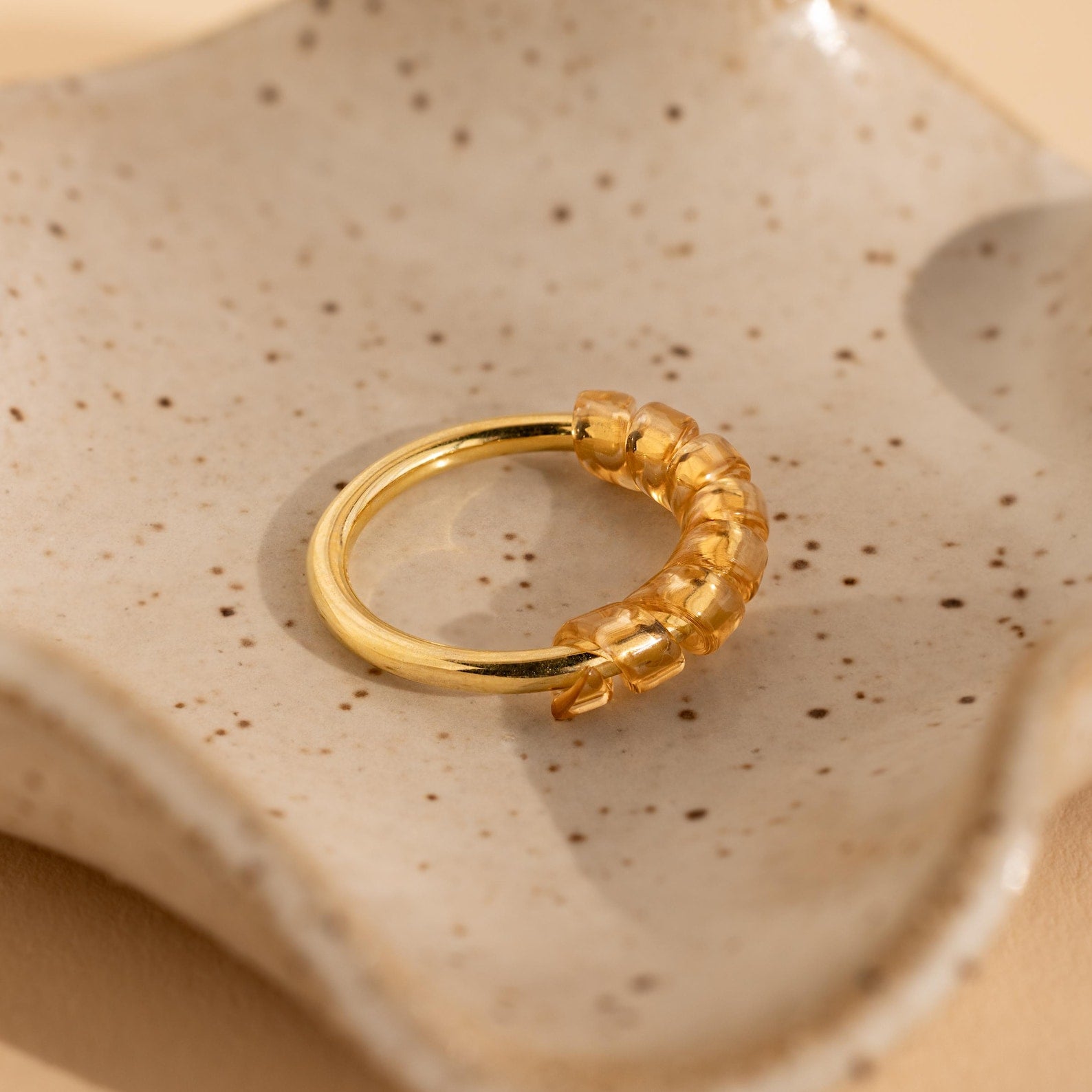 A gold ring with amber-colored beads, secured by a Ring Size Adjuster, rests on a speckled ceramic dish.