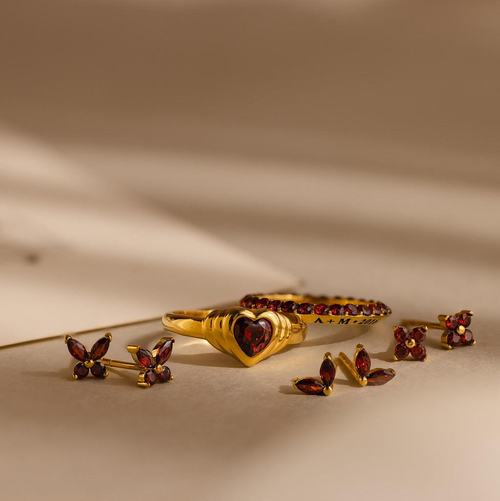 Gold and red gemstone earrings, such as Butterfly Birthstone Studs, and rings displayed on a beige surface with soft shadows.