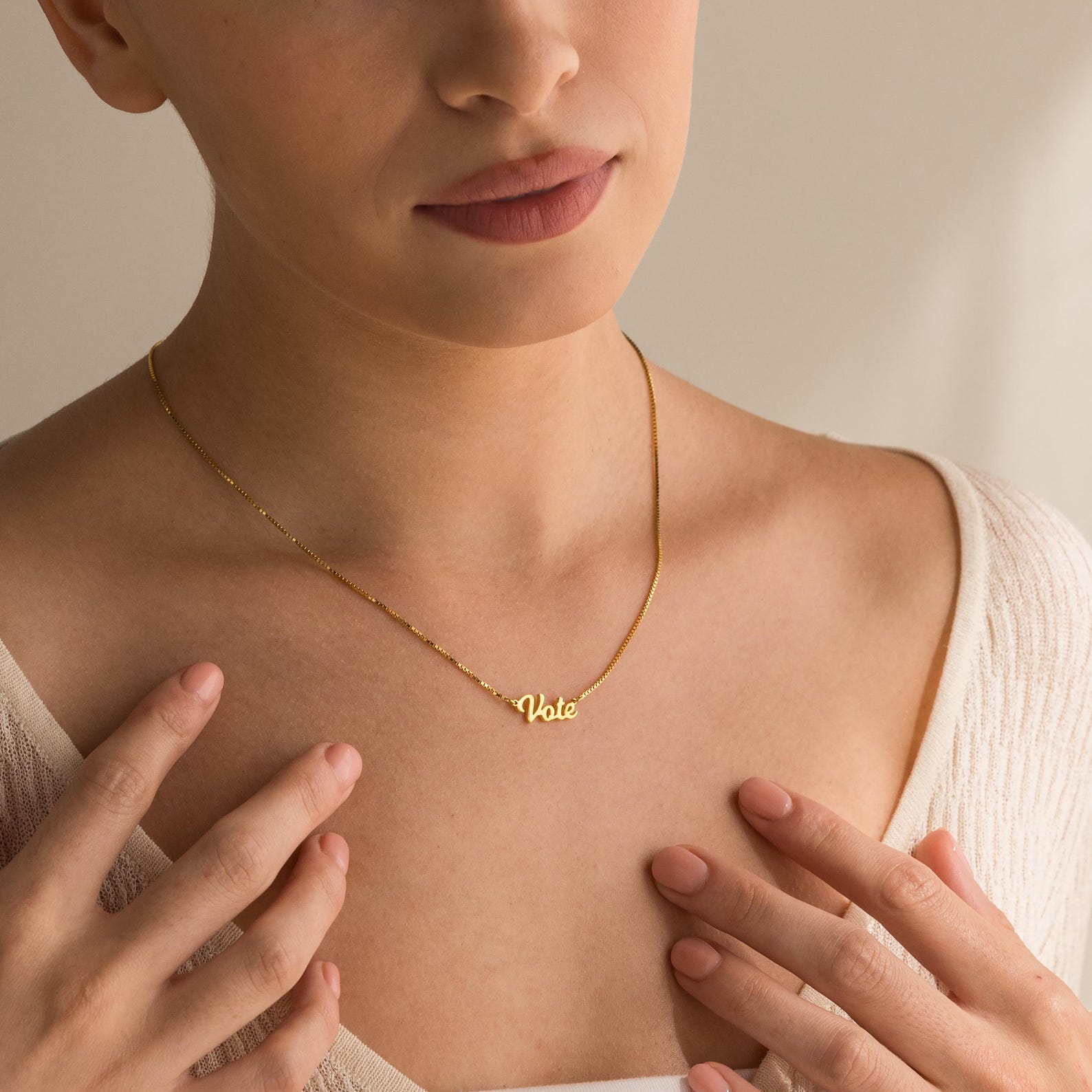 A woman wears an Essential Vote Necklace, holding the gold chain and pairing it with a light top—an inspiring gift for voters.