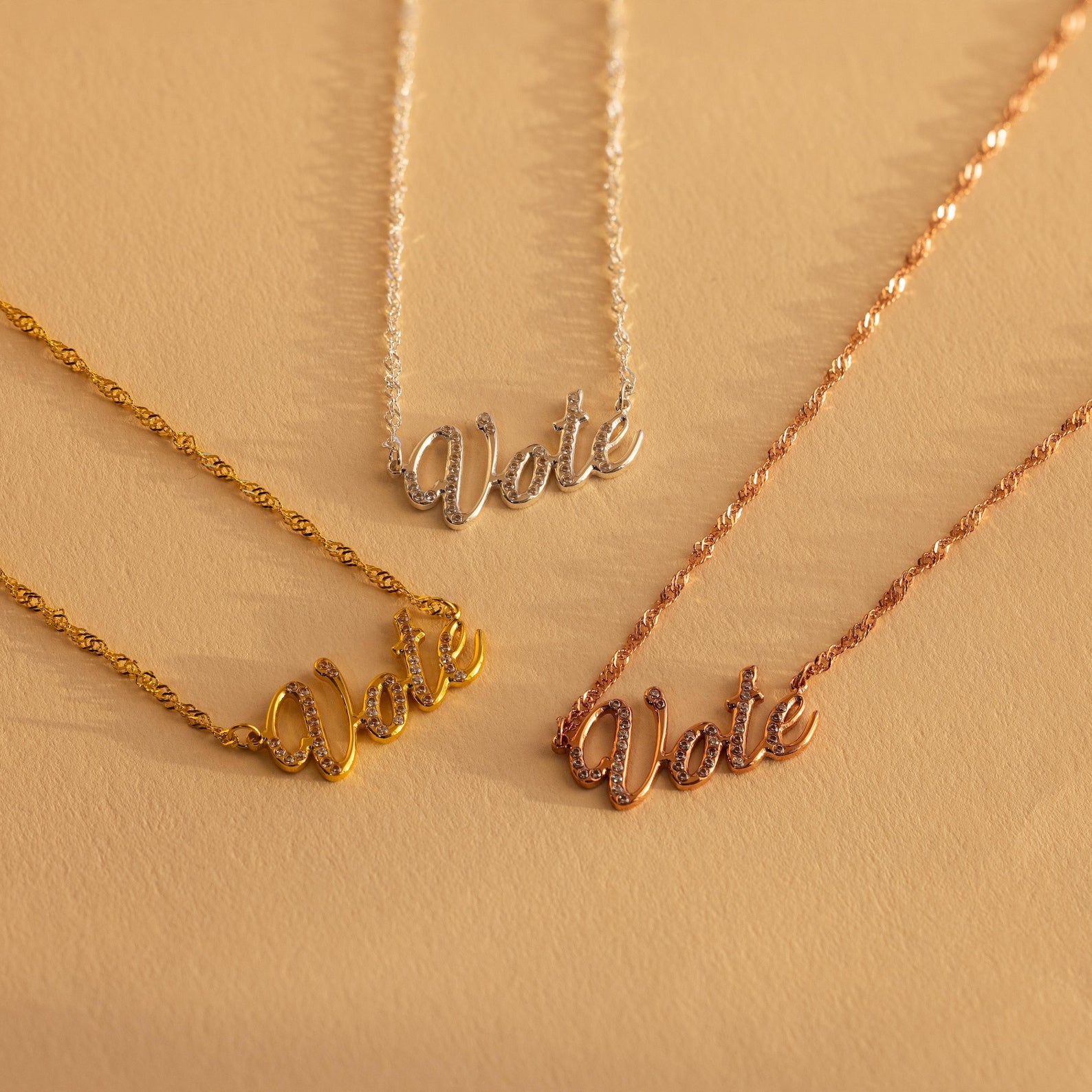 Three Essential Vote Necklaces in gold, silver, and rose gold rest on a beige surface—a meaningful and inspiring gift for voters or anyone who loves a symbolic voting necklace.