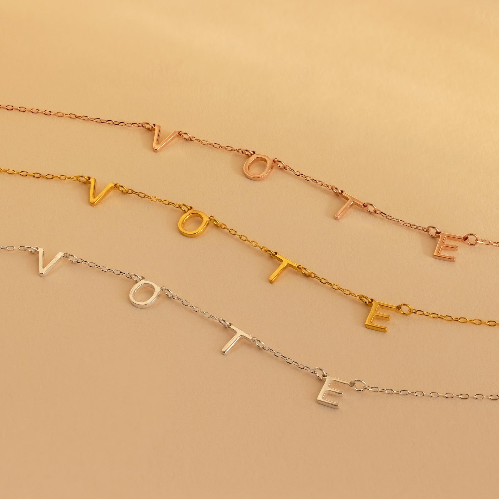 Three delicate Essential Vote Necklaces spell VOTE in gold, rose gold, and silver on a neutral background—a stylish gift for voters.