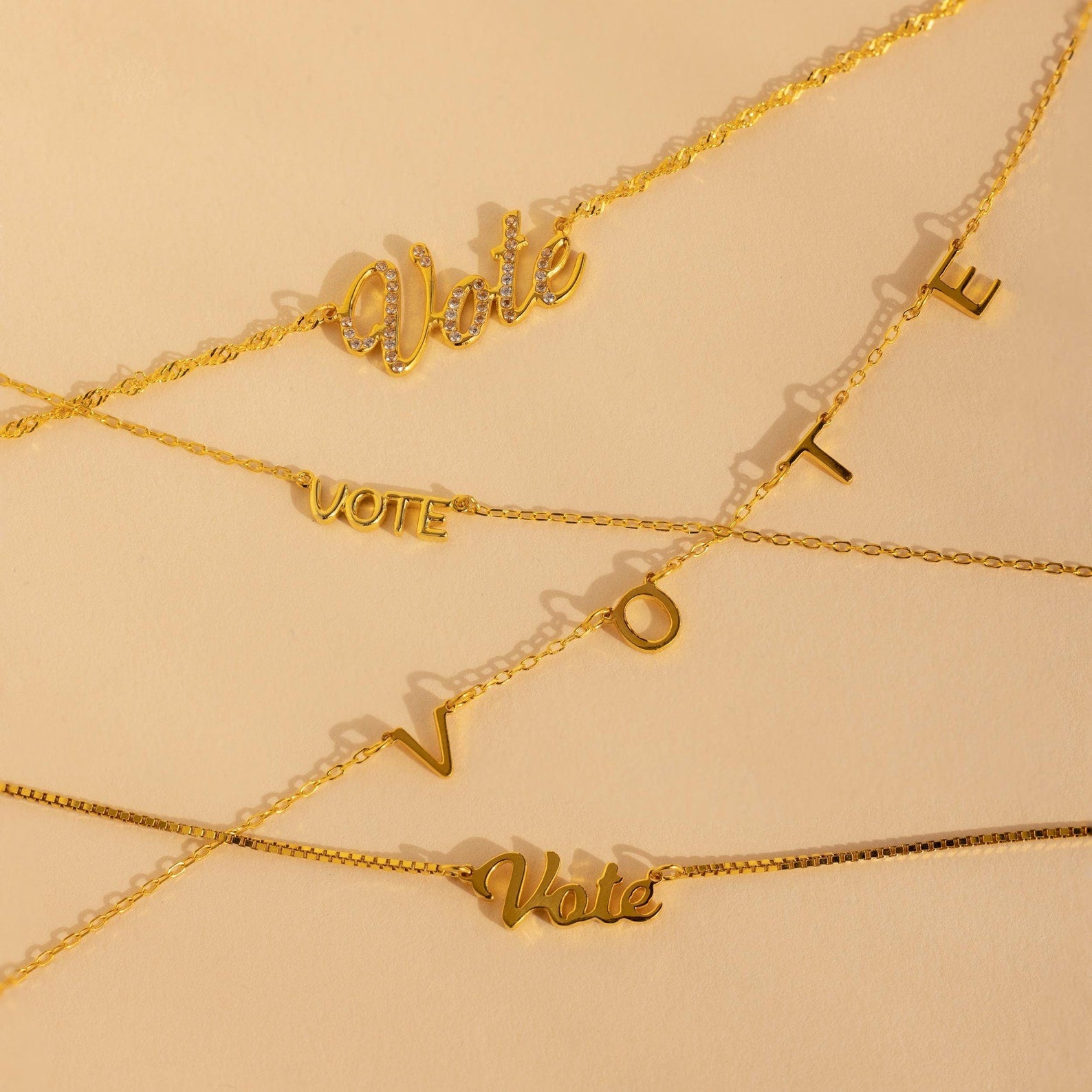 Essential Vote Necklaces: Four gold necklaces on a beige background, each with a unique “Vote” pendant—an ideal statement piece and thoughtful gift for voters.
