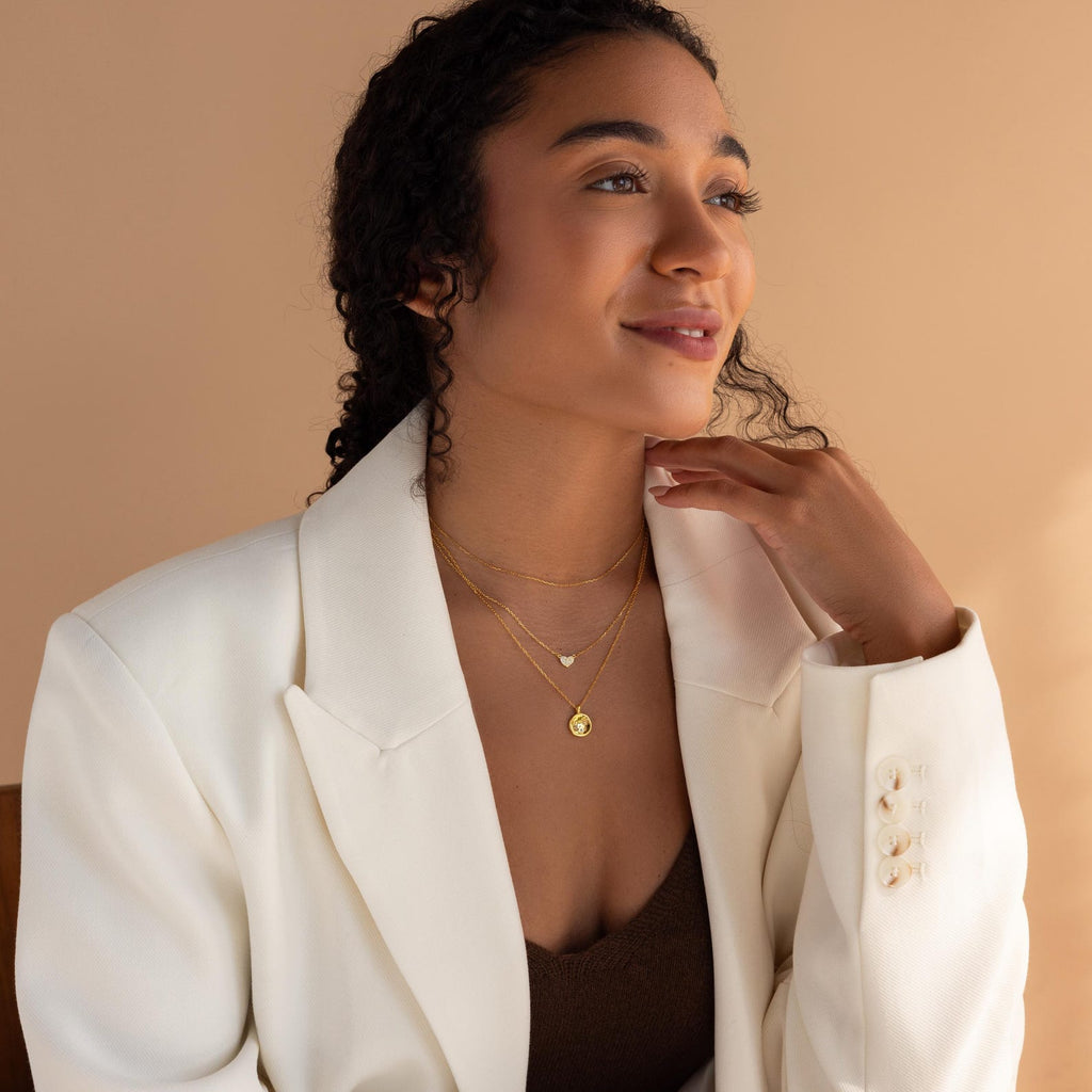 Woman in white blazer and layered gold necklaces, including the Baby Handprint Coin Necklace—an ideal gift for new moms—smiling as she looks to the side against a beige background.