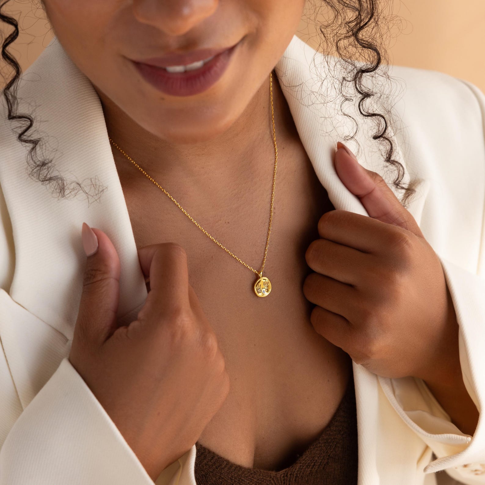 Smiling slightly, a woman in a white blazer wears the Baby Handprint Coin Necklace—a gold round pendant that's a perfect personalized keepsake and new mom gift.