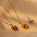 Three metallic Footprint Birthstone Necklaces with heart-shaped, multicolored gemstone pendants rest on beige fabric—an elegant personalized charm, ideal as parent and child jewelry.