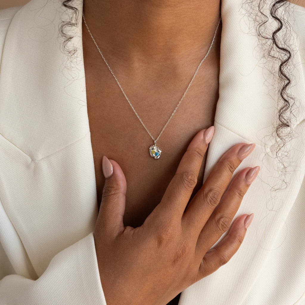 A person in a white blazer wears the Footprint Birthstone Necklace and has manicured nails touching their chest.