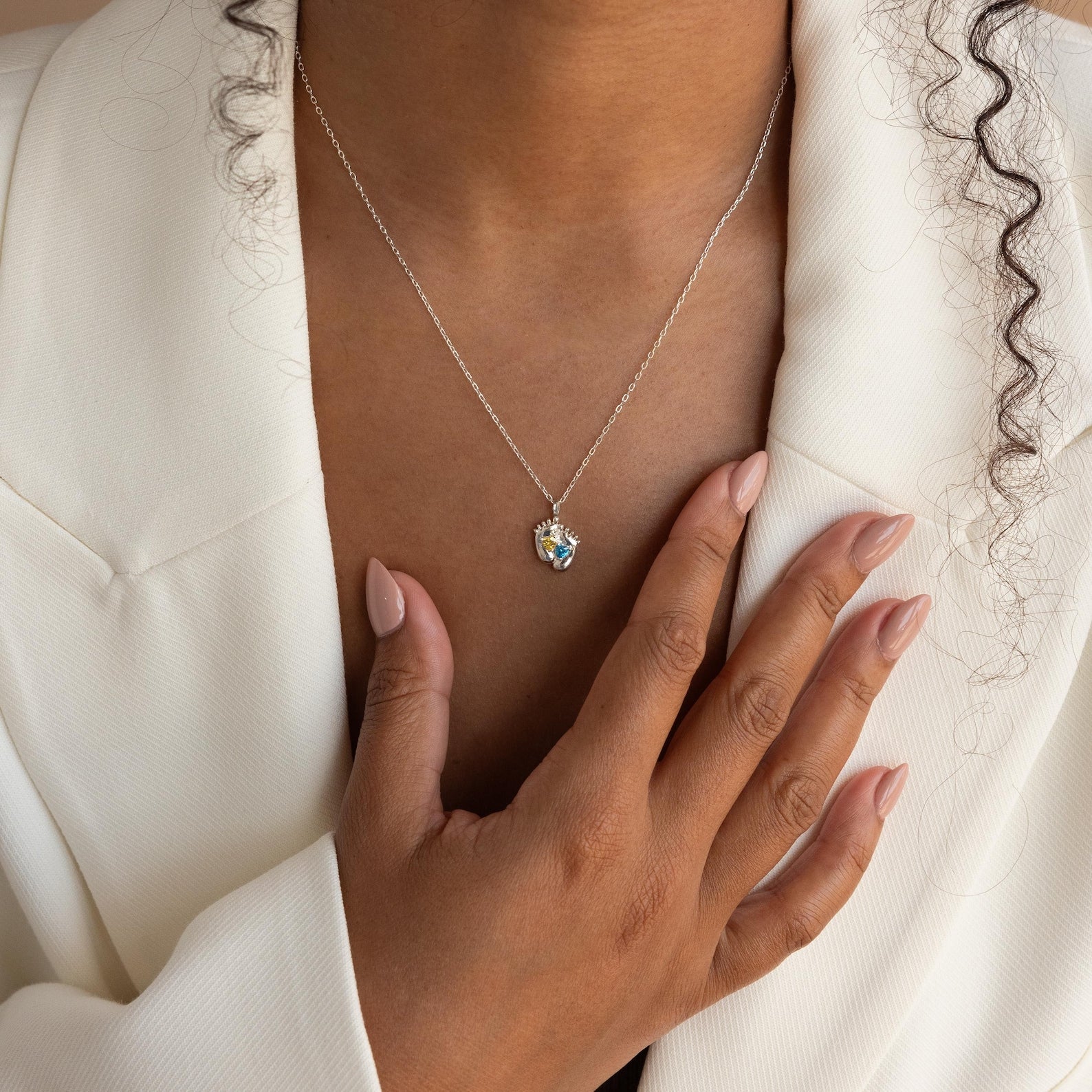 A person in a white blazer wears the Footprint Birthstone Necklace and has manicured nails touching their chest.