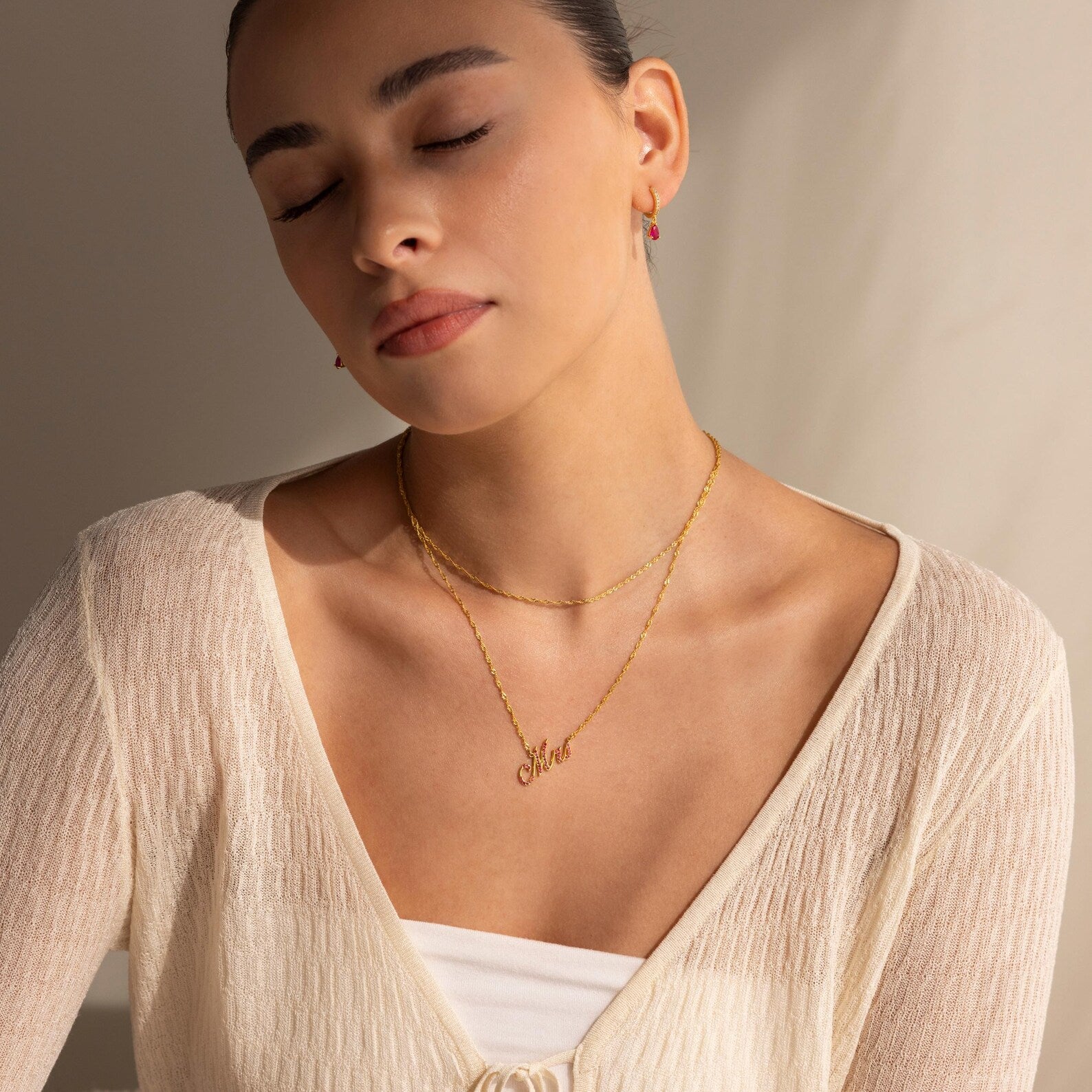 Woman with closed eyes, wearing an Elegant Birthstone Name Necklace and a cream knit top, sits in soft natural light.