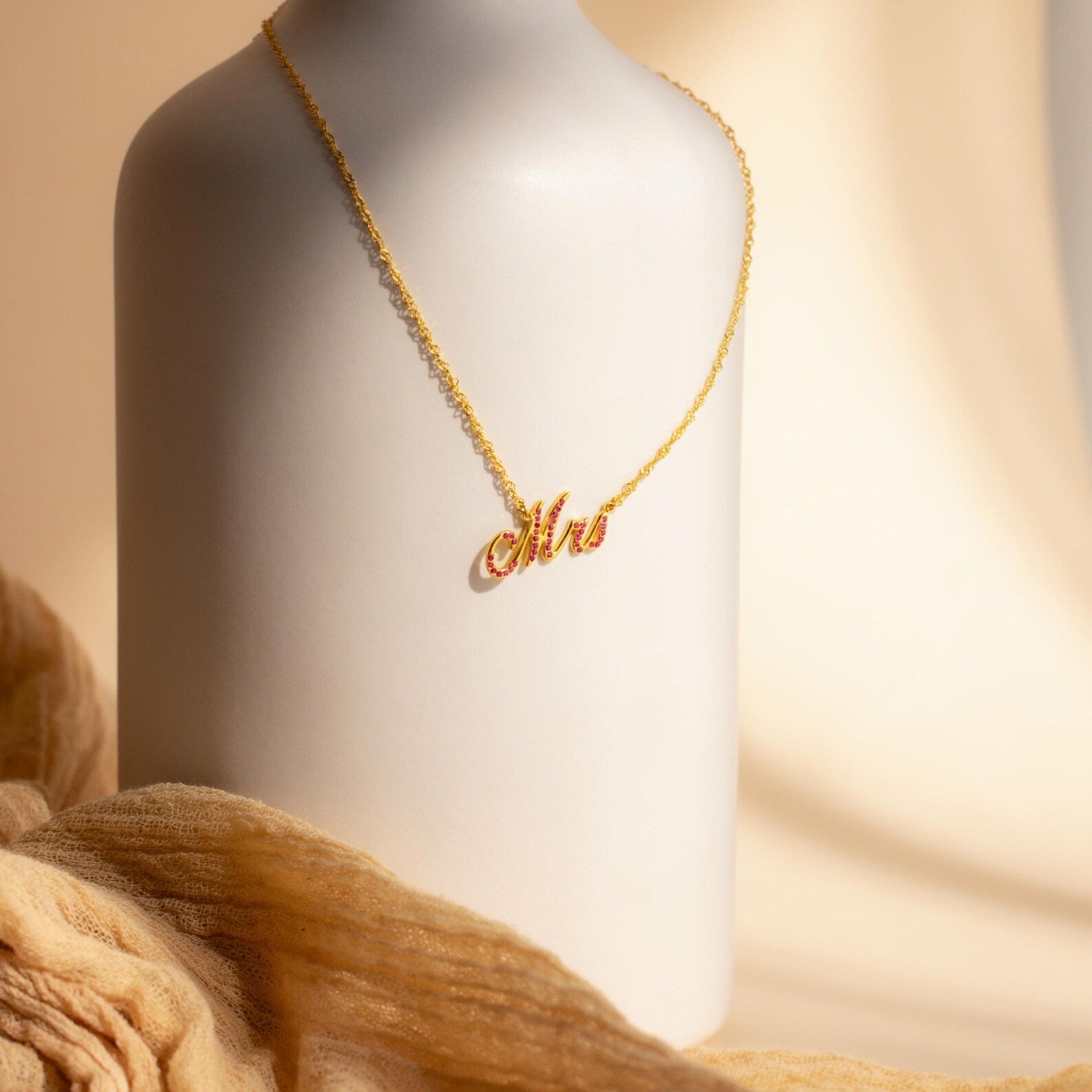 The Elegant Birthstone Name Necklace drapes over a smooth white vase with a soft beige fabric background, beautifully showcasing this custom jewelry piece with timeless elegance.