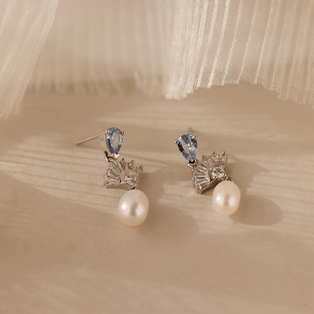 The Aquamarine Pearl Drop Earrings showcase blue gemstones, clear crystals, and dangling pearls, creating a vintage glamour look against a soft beige background.