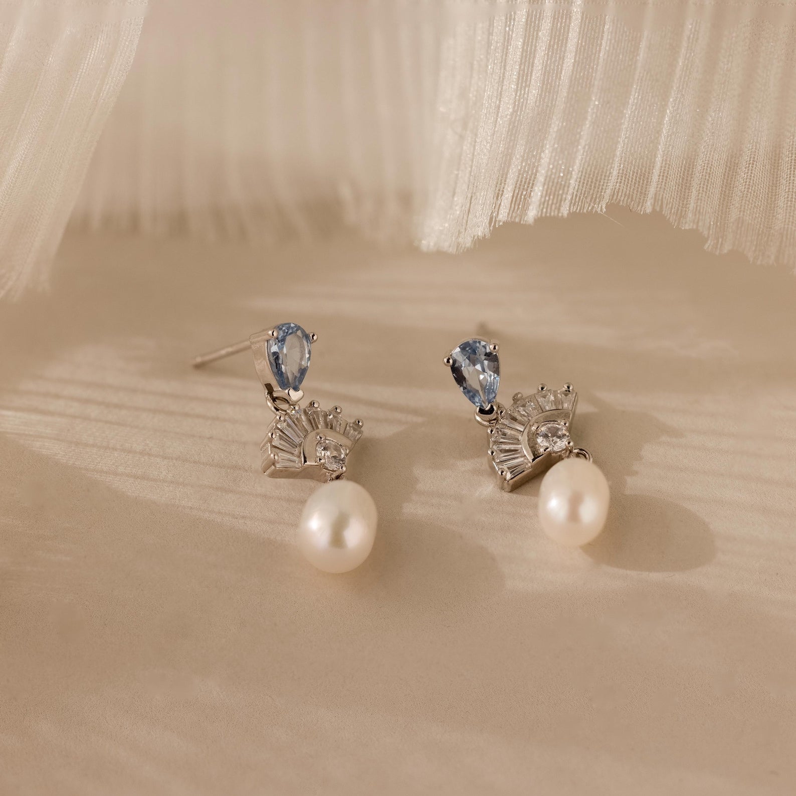 The Aquamarine Pearl Drop Earrings showcase blue gemstones, clear crystals, and dangling pearls, creating a vintage glamour look against a soft beige background.