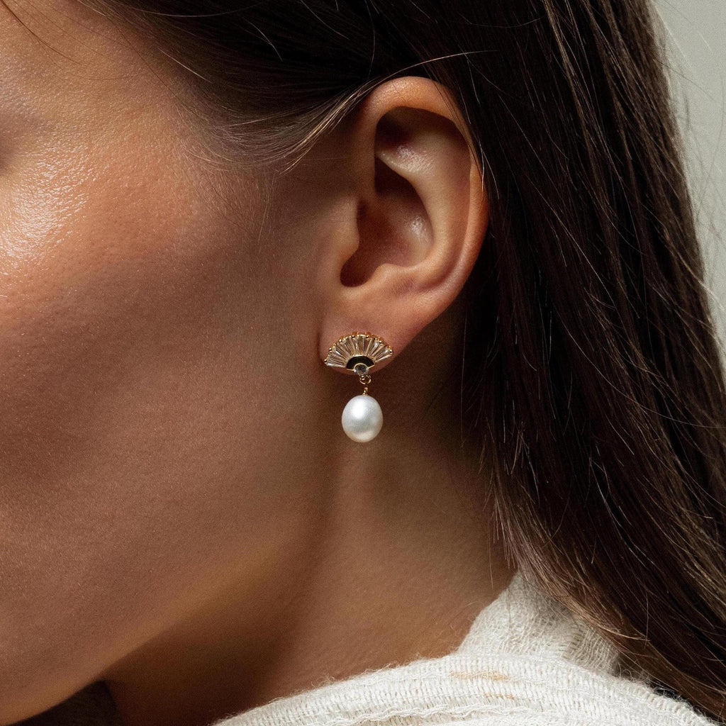 Close-up of a woman's ear wearing Art Deco Pearl Drop Earrings, featuring a gold fan-shaped design with a dangling pearl for vintage glamour.