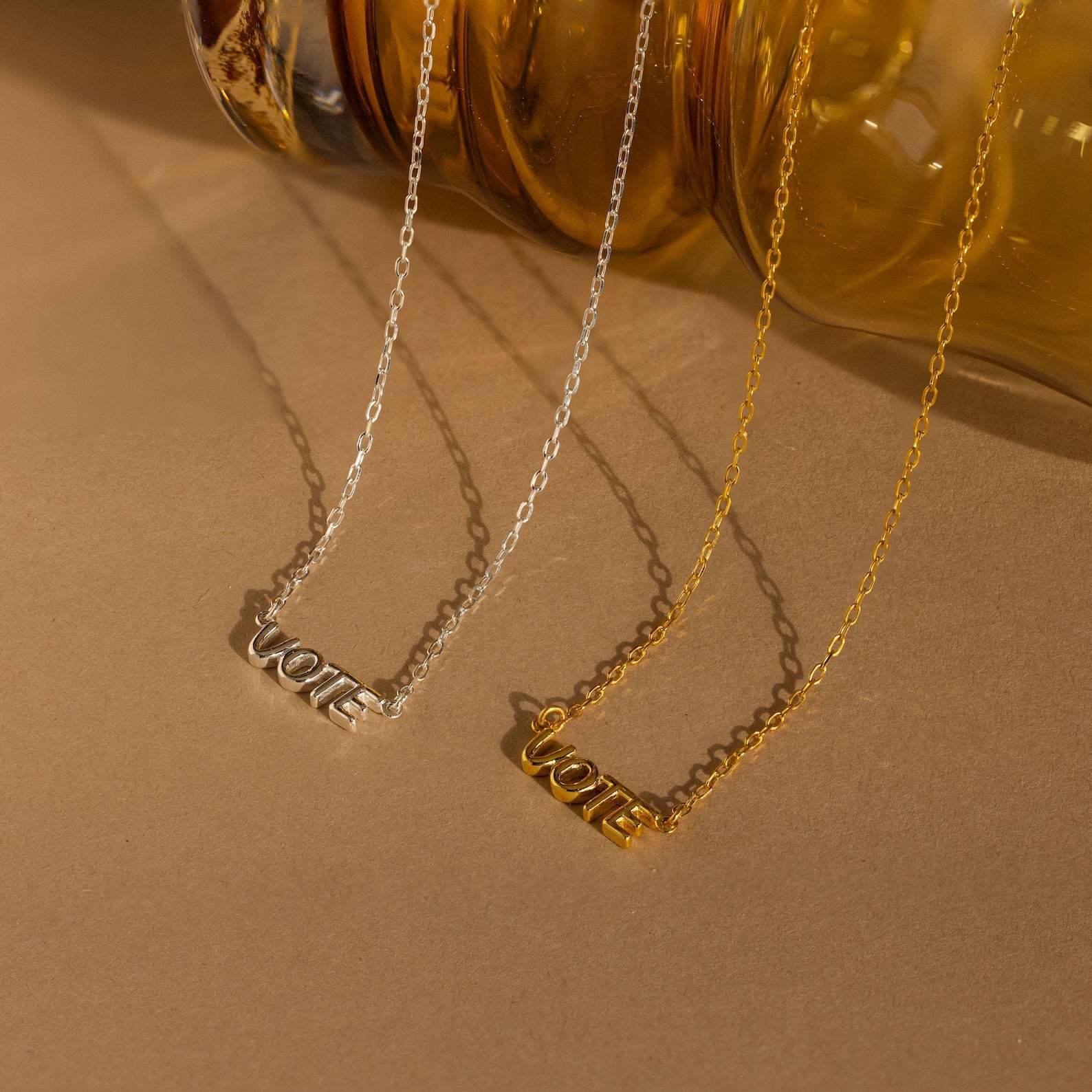 Essential Vote Necklaces in silver and gold with VOTE pendants are showcased on a tan surface beside a glass vase—an ideal gift for those looking to express their voice in style.