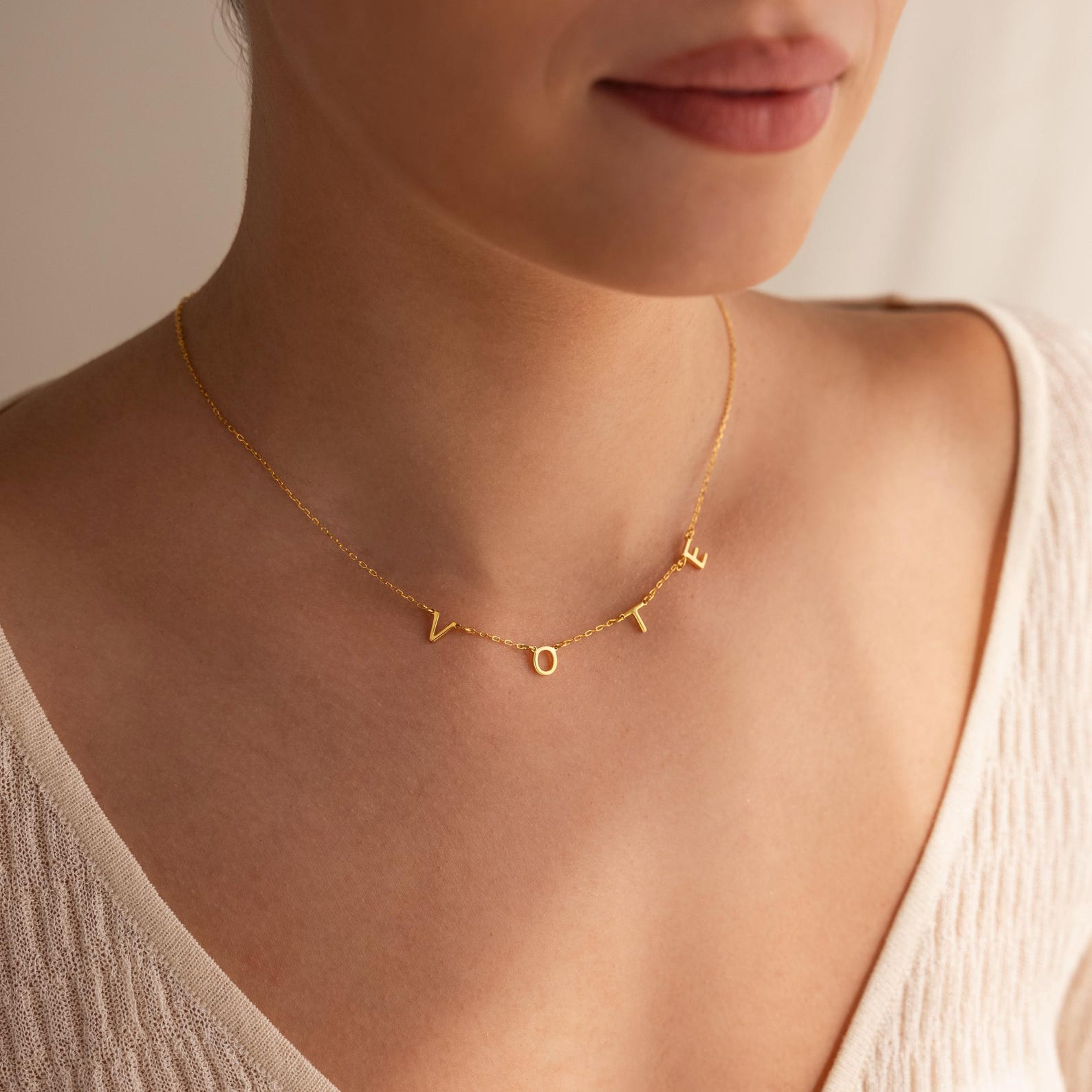 A woman wearing the Essential Vote Necklace with gold letter charms, styled with a cream knit top—a perfect voting accessory and thoughtful gift for voters.