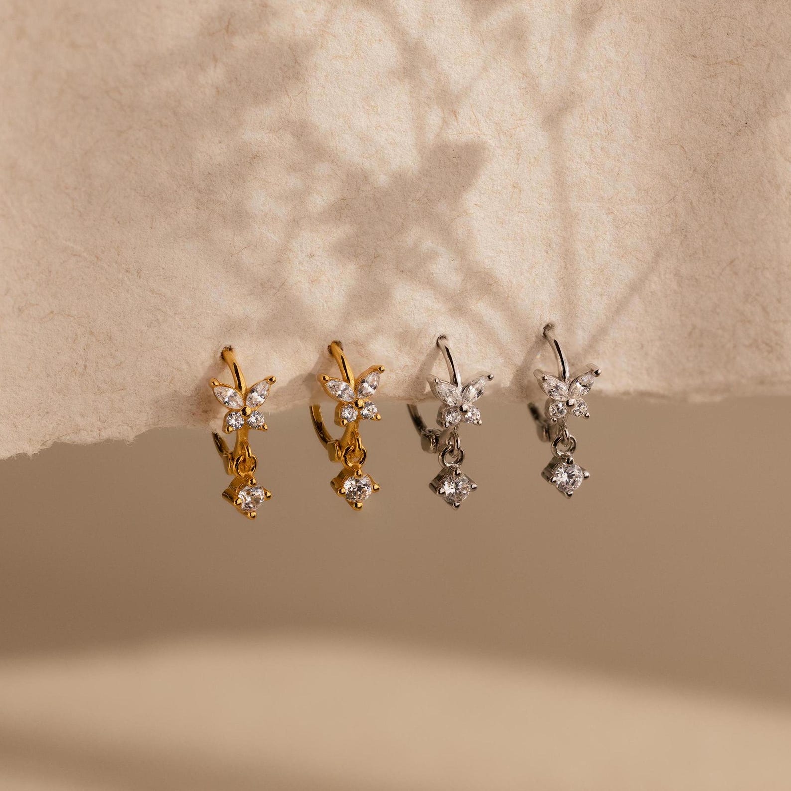Two pairs of dangling earrings, one gold and one silver, displayed on textured beige paper—perfect as a bridesmaid gift or to pair with your Diamond Butterfly Drop Jewelry Set in 18K Gold.