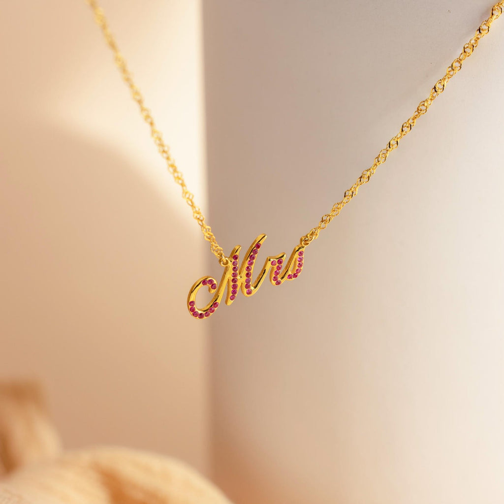 The Elegant Birthstone Name Necklace features a "Mrs" script pendant accented with pink stones, perfect as personalized or custom jewelry, showcased against a soft background.