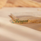 The Elegant Birthstone Name Necklace features “Morgan” in cursive with green stones on a beige surface—a stunning piece of personalized custom jewelry.