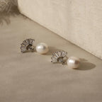 The Art Deco Pearl Drop Earrings, featuring elegant silver fan-shaped accents, are displayed on a beige surface and radiate vintage glamour.