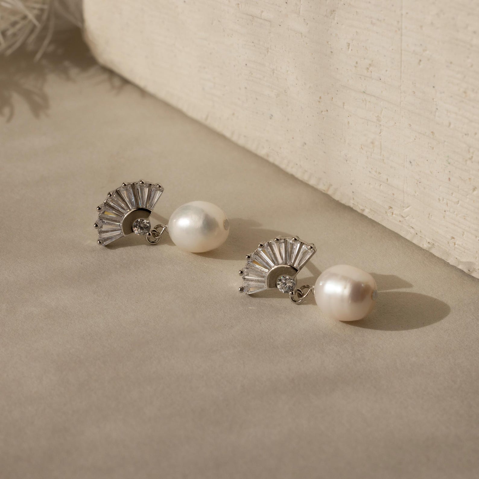 The Art Deco Pearl Drop Earrings, featuring elegant silver fan-shaped accents, are displayed on a beige surface and radiate vintage glamour.