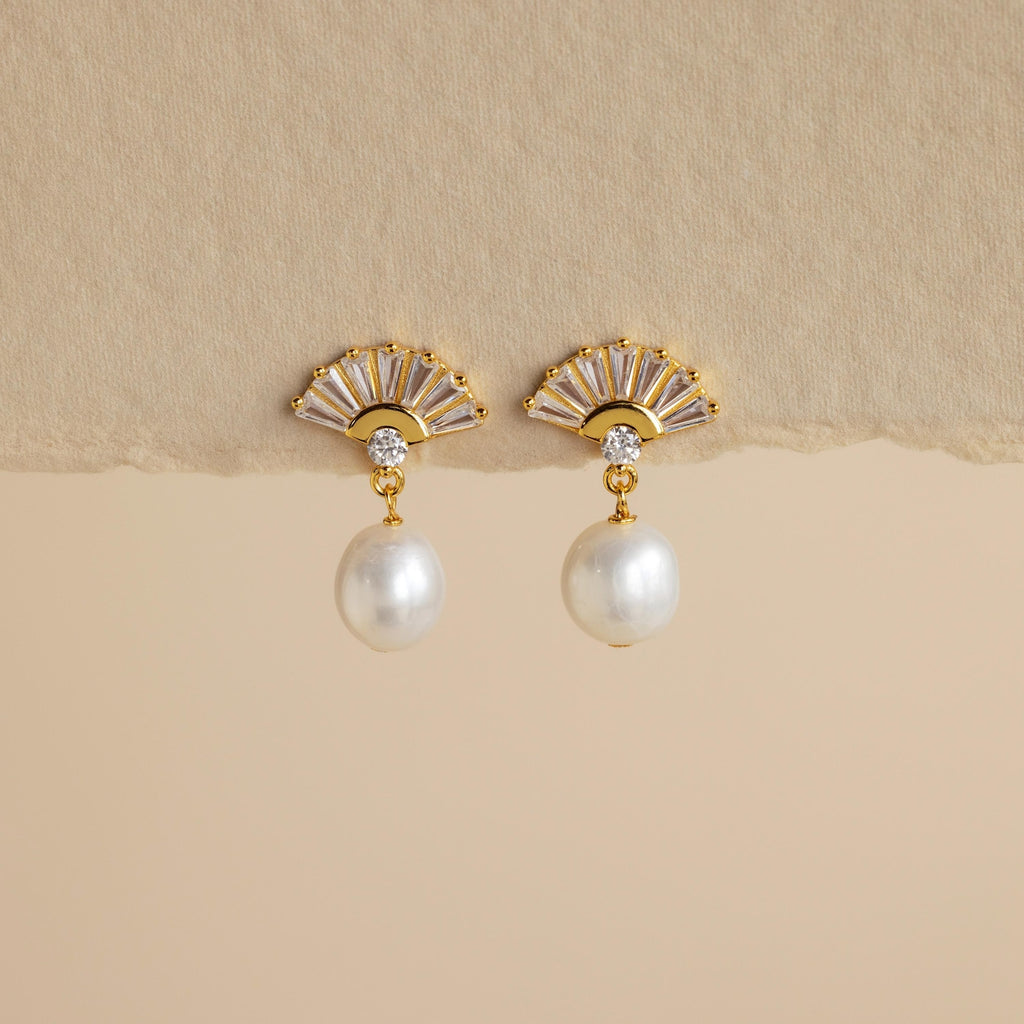 Art Deco Pearl Drop Earrings: Gold fan-shaped earrings with a small crystal and dangling white pearl, evoking vintage glamour. Set against a beige background.