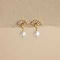 Art Deco Pearl Drop Earrings: Gold fan-shaped earrings with a small crystal and dangling white pearl, evoking vintage glamour. Set against a beige background.