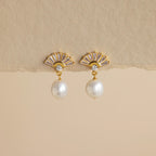 Art Deco Pearl Drop Earrings: Gold fan-shaped earrings with a small crystal and dangling white pearl, evoking vintage glamour. Set against a beige background.