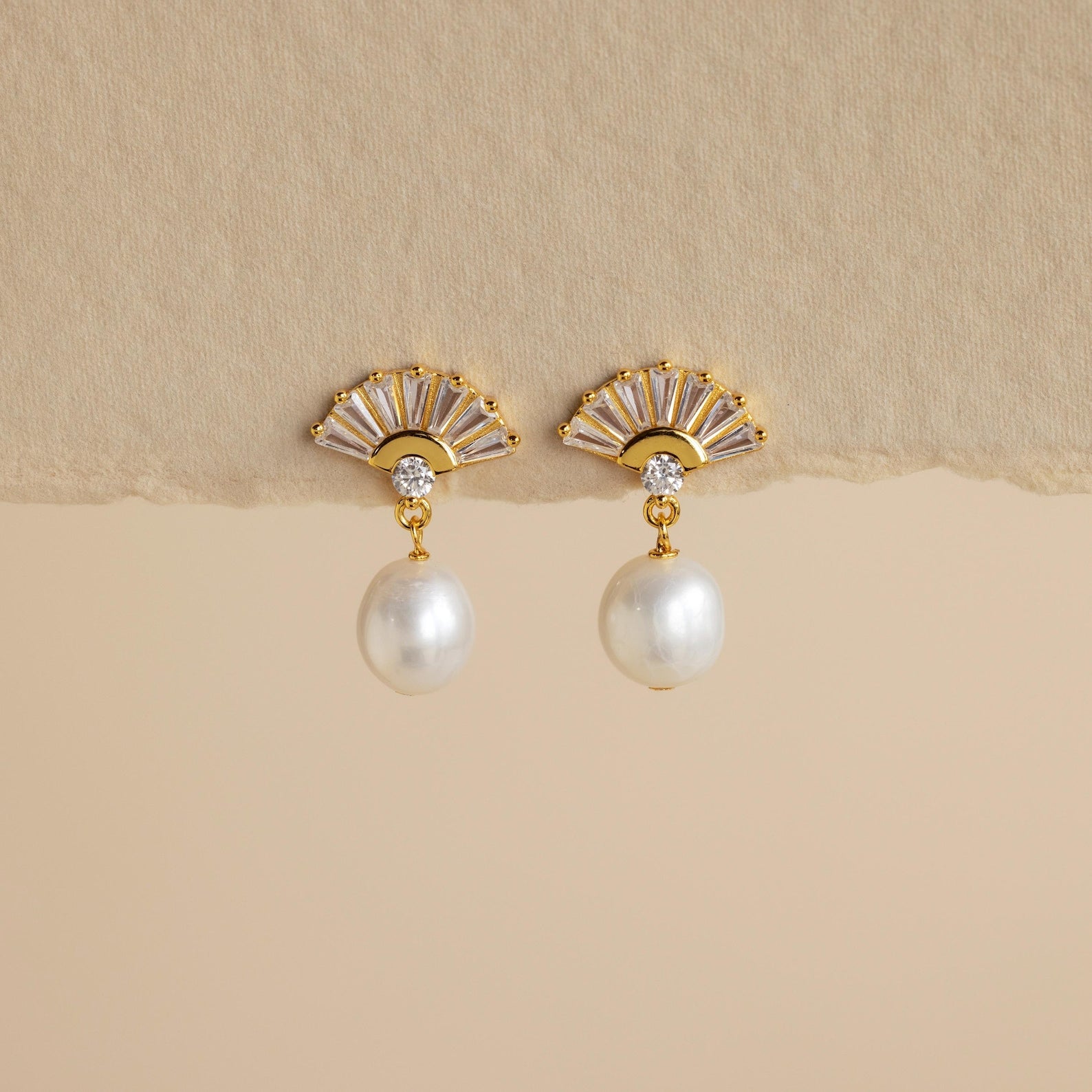 Art Deco Pearl Drop Earrings: Gold fan-shaped earrings with a small crystal and dangling white pearl, evoking vintage glamour. Set against a beige background.