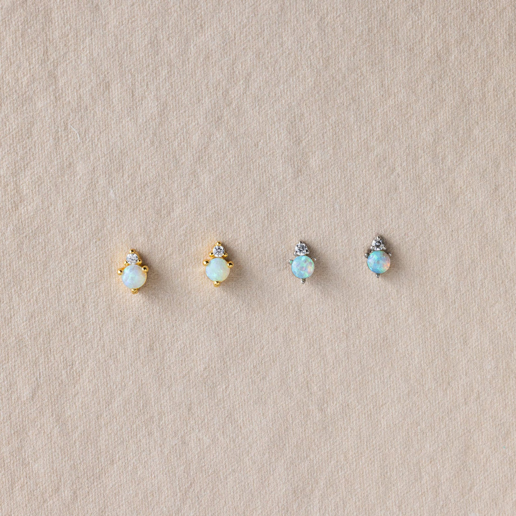 Four Raindrop Birthstone Studs—two gold and two silver, each with opal accents—are displayed on textured beige fabric. These dainty earrings offer a subtle touch of elegance to any look.