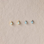 Four Raindrop Birthstone Studs—two gold and two silver, each with opal accents—are displayed on textured beige fabric. These dainty earrings offer a subtle touch of elegance to any look.
