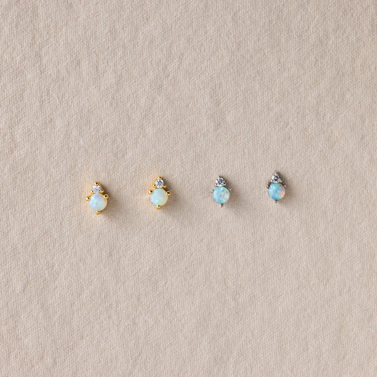 Four Raindrop Birthstone Studs—two gold and two silver, each with opal accents—are displayed on textured beige fabric. These dainty earrings offer a subtle touch of elegance to any look.