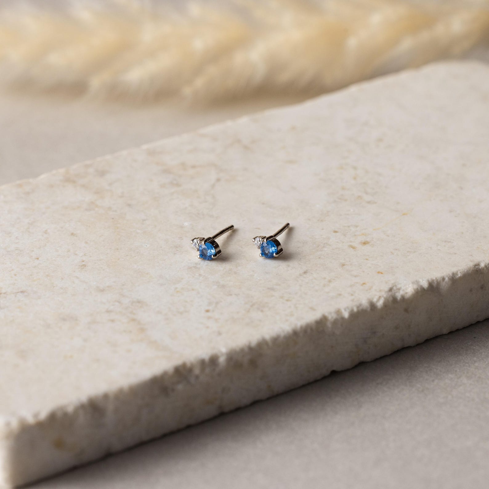 Raindrop Birthstone Studs: small, silver dainty earrings featuring blue gemstones, perfect as birthstone studs and displayed on a textured, light-colored stone surface.